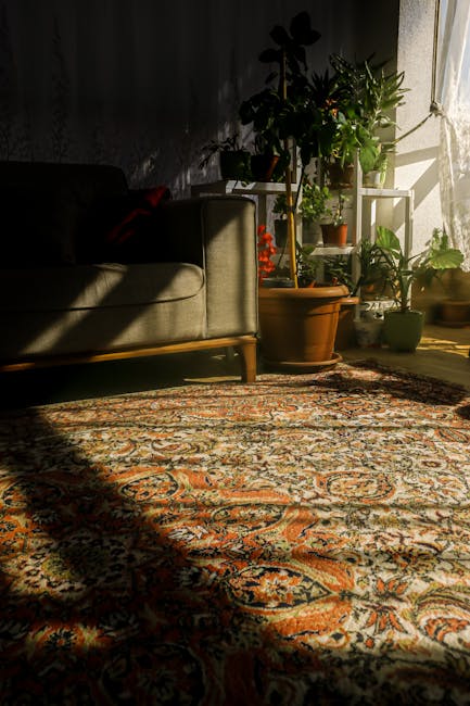 The image depicts a living room corner with a patterned Oriental-style carpet covering the wooden floor, illuminated by natural sunlight streaming through a nearby window. To the left, there is a beige upholstered sofa with wooden legs, displaying a subtle sheen on its fabric suggesting recent deep cleaning or proper maintenance. Behind the sofa, a collection of various houseplants in terracotta and ceramic pots are arranged on a white metal plant stand and directly on the floor, adding greenery to the space. The sunlight highlights the vibrant colors of the plants and casts soft shadows across the carpet, enhancing the room’s warm and inviting atmosphere. The scene emphasizes the importance of surface cleaning and front-line maintenance of household furnishings, aligning with tips on carpet and domestic cleaning provided by Harrow Carpet Cleaners on their page about carpet cleaning Harrow on the Hill period homes.