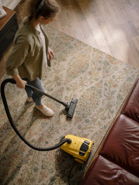 A woman using a yellow vacuum cleaner to perform deep surface cleaning on a patterned area rug, which features intricate floral and geometric designs in beige, orange, green, and blue tones, situated in a living room with wooden flooring and a red leather sofa. The scene is well-lit with natural light, highlighting the cleanliness of the carpet and surrounding wooden floor. The woman is dressed casually in a green jacket, white sneakers, and dark jeans. This image illustrates domestic cleaning practices, with a focus on carpet sanitisation and maintaining a tidy living space, as part of the services offered by Harrow Carpet Cleaners, aligning with tips for period home maintenance in Harrow on the Hill.