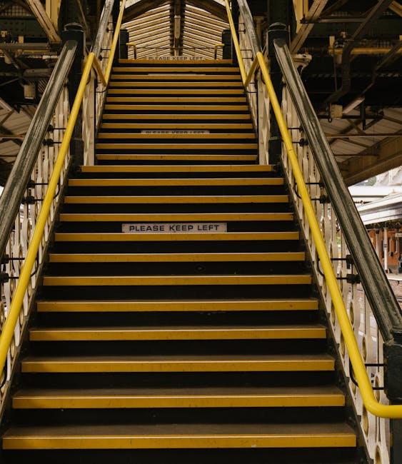 Image of a narrow industrial staircase with yellow and black striped safety markings on each step, leading up to a higher level within a building structure. The staircase has metal railings painted yellow, positioned on both sides, and features a sign on the steps that reads 'PLEASE KEEP LEFT'. The surrounding environment includes metal beams and industrial equipment, with lighting casting shadows on the steps. This setting emphasizes the importance of maintaining cleanliness and safety in challenging access areas, reflecting the expertise of Harrow Carpet Cleaners in thorough surface cleaning and sanitation for commercial or residential spaces, especially in hard-to-reach locations such as narrow stairs in flats or industrial buildings.