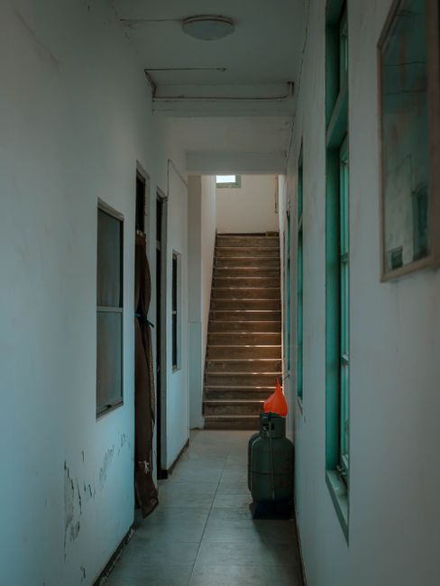 A narrow indoor staircase with wooden steps leading upwards, situated in a confined corridor with white painted walls. The corridor has a tiled floor and is illuminated by natural light coming from a small window at the top of the stairs. On the right side, there are large glass windows with visible green frames, allowing additional daylight into the space. Along the left wall, there are two small, open-fronted service or storage compartments, and a brown cloth or bag hangs nearby. Near the base of the stairs, an orange safety cone and a black cleaning or storage container are placed, suggesting ongoing maintenance or cleaning activities. The surfaces appear clean but show some signs of wear and minor dirt, typical of a residential or commercial property in need of regular surface cleaning and sanitisation. This image is associated with Harrow Carpet Cleaners' specialist services for cleaning difficult-to-access areas, such as narrow stairs, ensuring hygiene and cleanliness in residential and commercial properties.