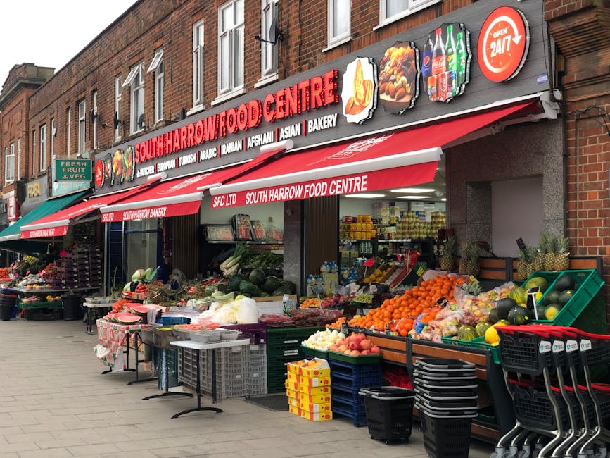 The exterior view of South Harrow Food Centre located in Harrow town centre features a brick building with a prominent red and white signage displaying the centre's name and various food types such as Asian, Afghan, Turkish, and bakery items. The entrance is shaded by a red awning, under which fresh produce like fruits and vegetables are displayed on tables and green crates along the sidewalk. To the left, additional produce is arranged near a small green stand, with an assortment of colorful fruits and vegetables including oranges, apples, and watermelons. The pavement in front of the store is clean, and the scene is well-lit with natural daylight, highlighting the vibrant colors of the fresh goods and the tidy arrangement of the displays, indicating a maintained and hygienic retail environment. This image exemplifies the importance of surface cleaning and sanitation in retail spaces, relevant to the detailed cleaning guide for Harrow town centre flats provided by Harrow Carpet Cleaners.