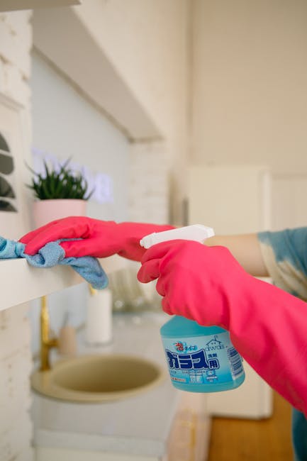 Close-up of a person wearing bright pink rubber gloves using a spray bottle of cleaning solution while wiping a white shelf in a kitchen. The surface appears to be a smooth, painted or laminated shelf with a small potted green plant and a blue cloth placed nearby. In the background, there is a beige or light-colored brick wall, a sink, and other kitchen fixtures, all appearing clean and well-maintained. The scene depicts domestic surface cleaning focused on sanitisation and hygiene, aligning with professional end of tenancy cleaning services promoted by Harrow Carpet Cleaners for properties like flats in Harrow town centre.