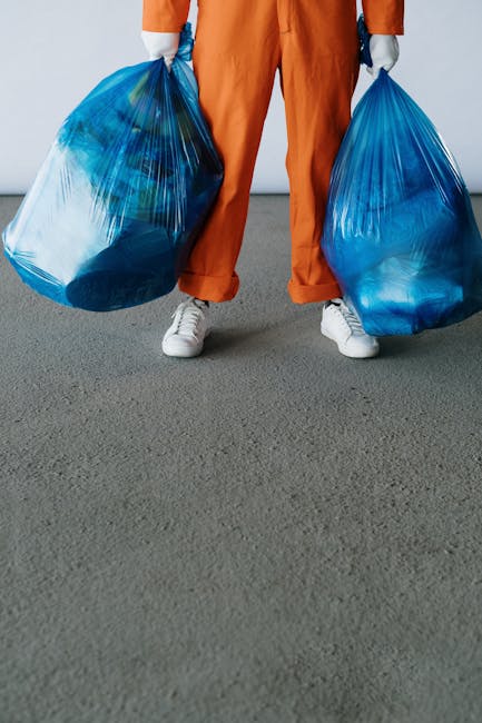 A person's hand holding a blue plastic waste bag, positioned against a plain white background. The bag appears to be partially filled and is crinkled, indicating it has been used for waste collection.