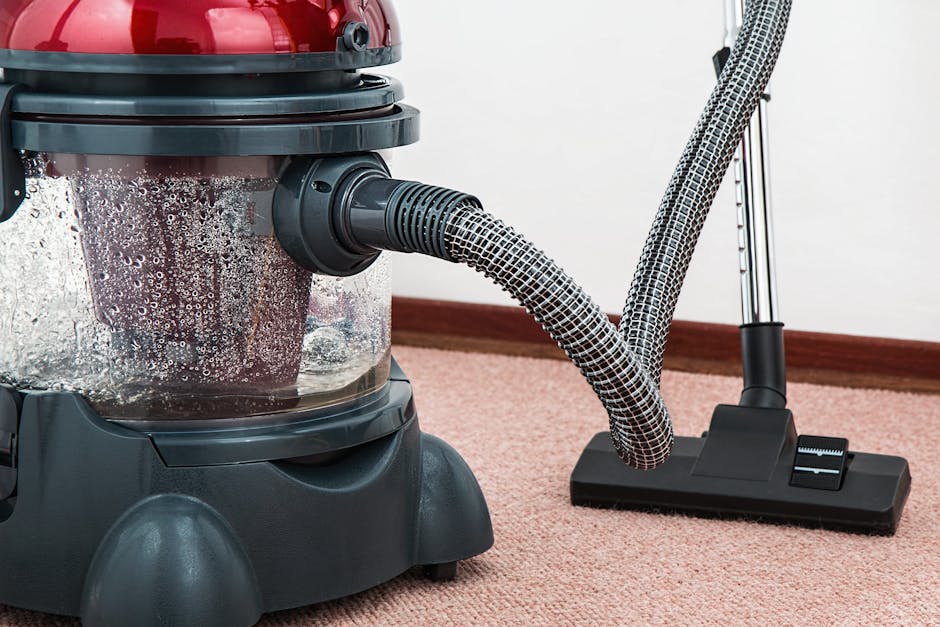 A close-up view of a vacuum cleaner with a red top and transparent dust container, positioned on a beige carpeted floor in a residential setting. The vacuum hose, covered with a black and white patterned cloth, is connected to the device and extends towards the carpet. The room features a white wall with a wooden baseboard, and the scene is well-lit, emphasizing the cleanliness and maintenance of the carpet surface, indicative of professional deep cleaning services by Harrow Carpet Cleaners in accordance with hygiene and surface cleaning practices.