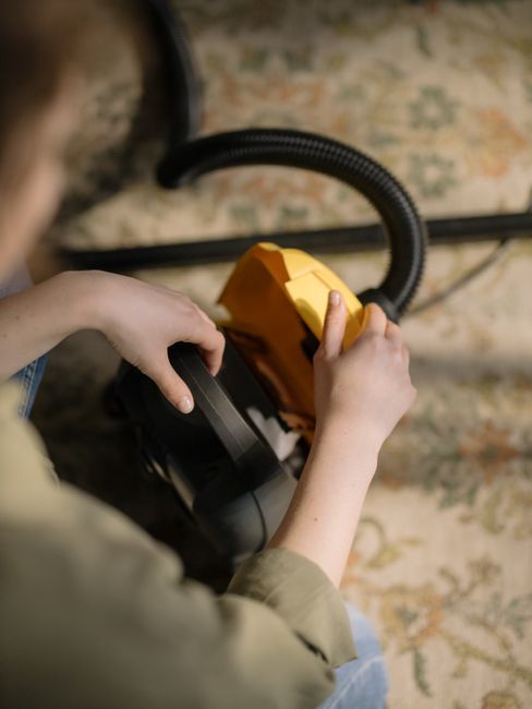 Close-up view of a person holding and operating a yellow and black vacuum cleaner, cleaning a patterned area rug in a well-lit room. The person's hands are visible, with one hand on the vacuum handle and the other adjusting the cord or attachment. The rug features an intricate floral design with muted tones, and the surrounding surface appears clean. The scene suggests surface cleaning or deep cleaning as part of maintaining hygiene and cleanliness in a domestic setting. The background hints at a room with natural light, emphasizing the importance of effective cleaning to avoid hidden charges in Harrow carpet cleaning. Harrow Carpet Cleaners specializes in professional cleaning services that focus on thorough sanitisation and care for various surfaces.}