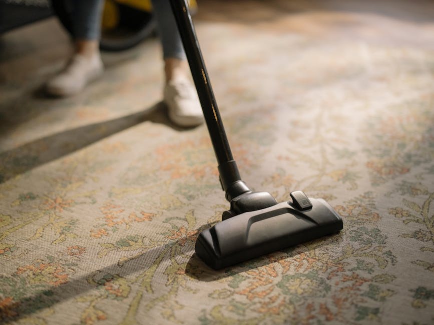 A close-up view of a vacuum cleaner with a red top and transparent dust container, positioned on a beige carpeted floor in a residential setting. The vacuum hose, covered with a black and white patterned cloth, is connected to the device and extends towards the carpet. The room features a white wall with a wooden baseboard, and the scene is well-lit, emphasizing the cleanliness and maintenance of the carpet surface, indicative of professional deep cleaning services by Harrow Carpet Cleaners in accordance with hygiene and surface cleaning practices.