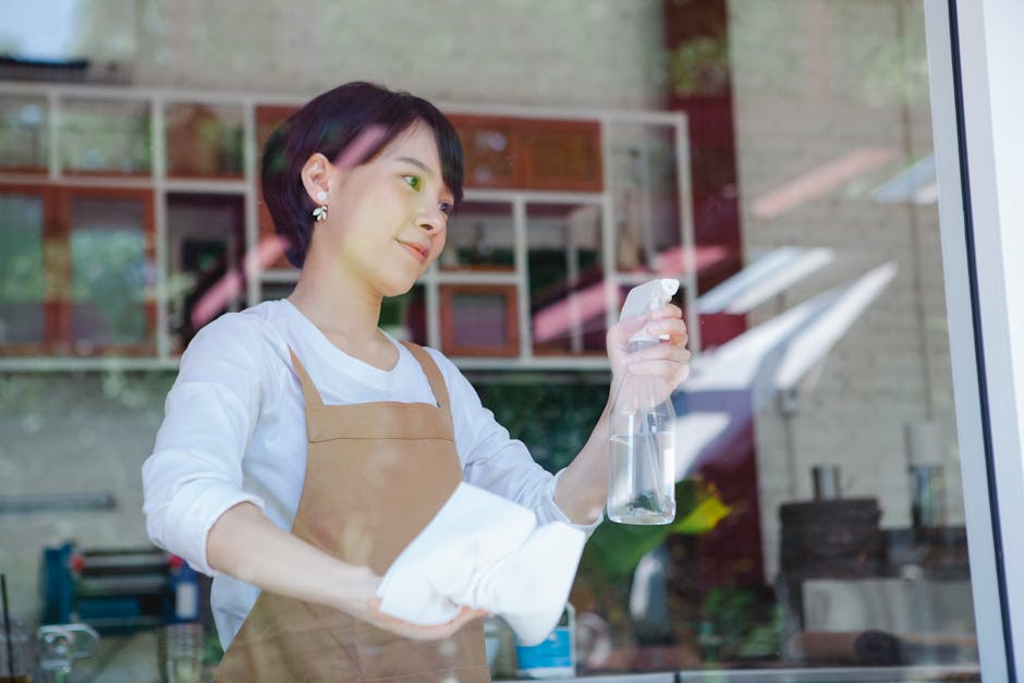 A female cleaning professional wearing a white shirt and light brown apron is standing inside a commercial kitchen, seen through a glass window. She is holding a spray bottle of cleaning solution in h
