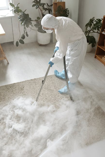 A professional cleaner dressed in white protective suits, gloves, and a face mask is performing deep surface cleaning on a light beige carpet in a bright living room. The cleaner is using a steam clea