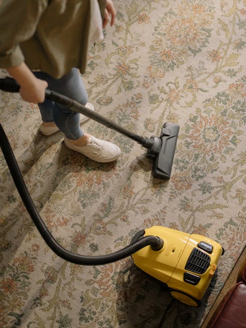 A professional cleaner dressed in white protective suits, gloves, and a face mask is performing deep surface cleaning on a light beige carpet in a bright living room. The cleaner is using a steam clea