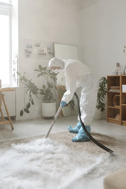 A professional worker dressed in a white protective suit, gloves, and a face mask is using a vacuum or steam cleaning equipment to deep clean a plush, beige carpet in a well-lit, modern living room. The room features white painted walls, a small wooden shelving unit with decorative items, and potted houseplants near a large window that allows natural light to brighten the space. The flooring is light-colored, and the overall scene emphasizes thorough surface cleaning and sanitary maintenance, as performed by Harrow Carpet Cleaners, aligning with the focus on same-day carpet cleaning and surface hygiene.