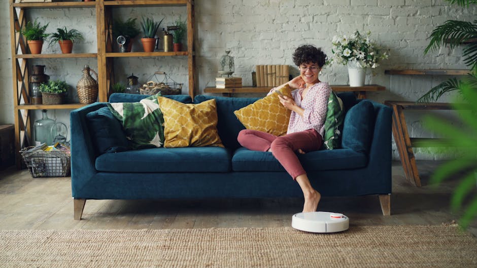 A woman sitting on a dark blue fabric sofa in a modern, well-lit living room, smiling while looking at her phone. The sofa is decorated with colorful throw pillows in green, yellow, and white, and is positioned against a white brick wall with wooden shelving filled with plants, decorative items, and books. A white robotic vacuum cleaner is on the wooden floor in front of the sofa, indicating domestic surface cleaning and maintenance. The room features natural light, a neutral-toned rug, and fresh indoor plants, creating a clean and inviting atmosphere that aligns with deep cleaning and sanitisation standards promoted by Harrow Carpet Cleaners.