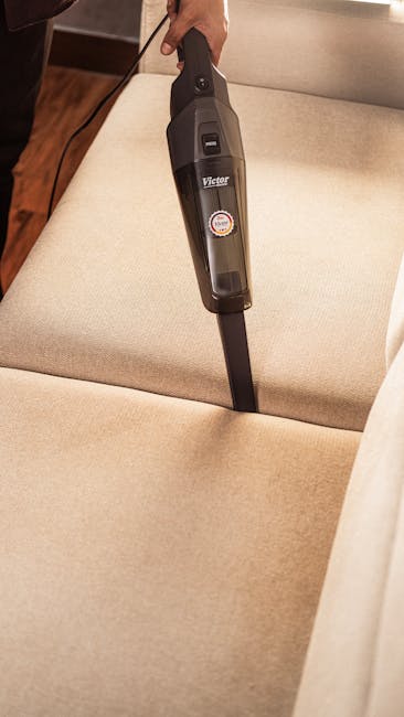 A close-up of a person using a handheld vacuum cleaner to clean the surface of a green upholstered sofa in a living room setting. The sofa has a fabric material with a tufted backrest and cushion, positioned against a white wall. In the background, part of a dark grey fabric sofa is visible, indicating a multi-piece seating arrangement. The room is well-lit with natural or artificial light, highlighting the clean and maintained appearance of the furniture. The scene reflects professional surface cleaning and maintenance, consistent with domestic cleaning services offered by Harrow Carpet Cleaners near North Harrow station.